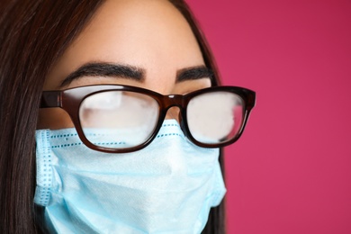 Photo of Young woman with foggy glasses caused by wearing disposable mask on pink background, closeup. Protective measure during coronavirus pandemic