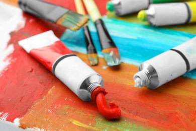 Photo of Tubes of colorful oil paints and brushes on canvas with abstract painting, closeup