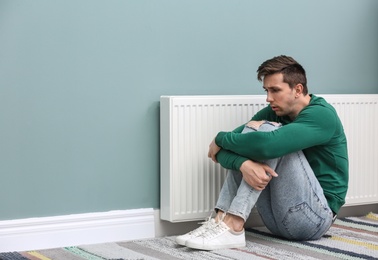 Photo of Sad young man suffering from cold on floor near radiator