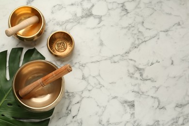 Golden singing bowls, mallets and monstera leaf on white marble table, flat lay. Space for text 