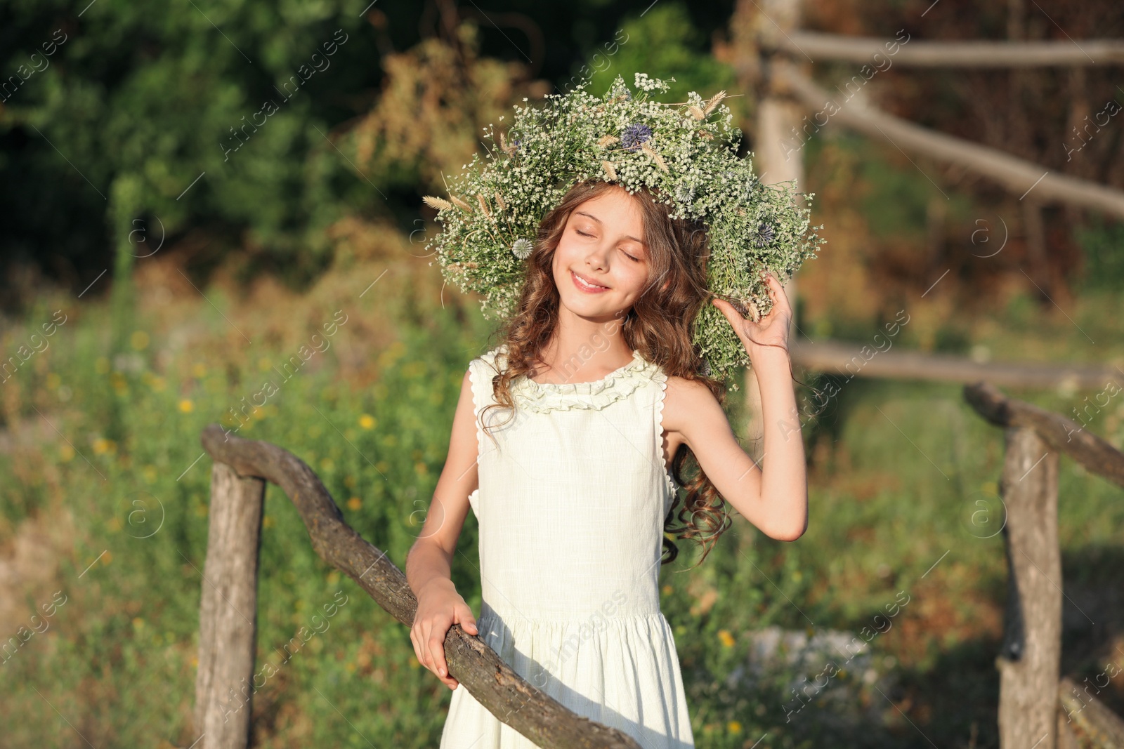 Photo of Cute little girl wearing wreath made of beautiful flowers near wooden fence outdoors on sunny day