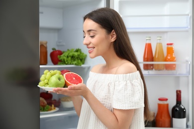 Photo of Concept of choice between healthy and junk food. Woman holding plate with fruits near refrigerator in kitchen