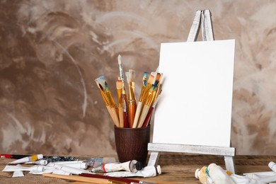 Photo of Many brushes, easel with blank canvas and paints on wooden table
