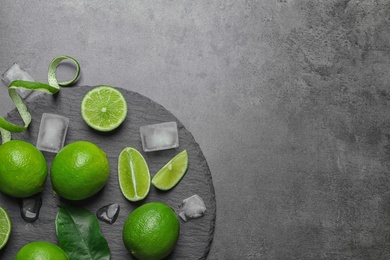 Slate plate with fresh ripe limes and ice cubes on gray background, top view