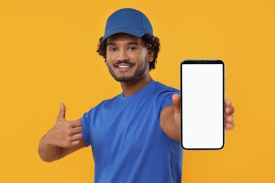 Happy courier holding smartphone with empty screen and showing thumbs up on orange background