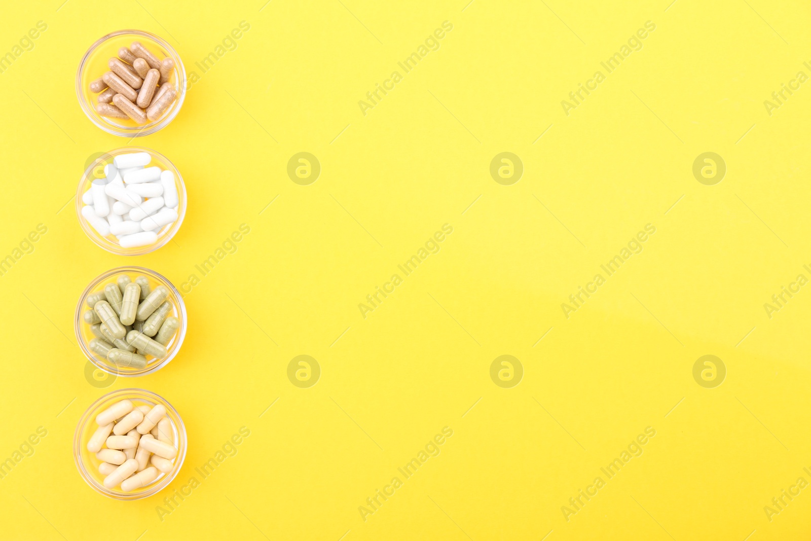 Photo of Different vitamin capsules in glass bowls on yellow background, flat lay. Space for text