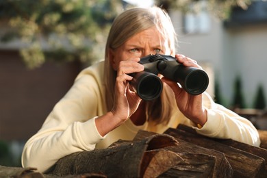 Concept of private life. Curious senior woman with binoculars spying on neighbours over firewood outdoors