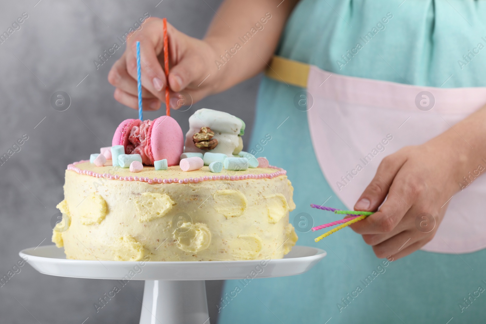 Photo of Woman putting candles on cake decorated with macarons and marshmallows against grey background, closeup