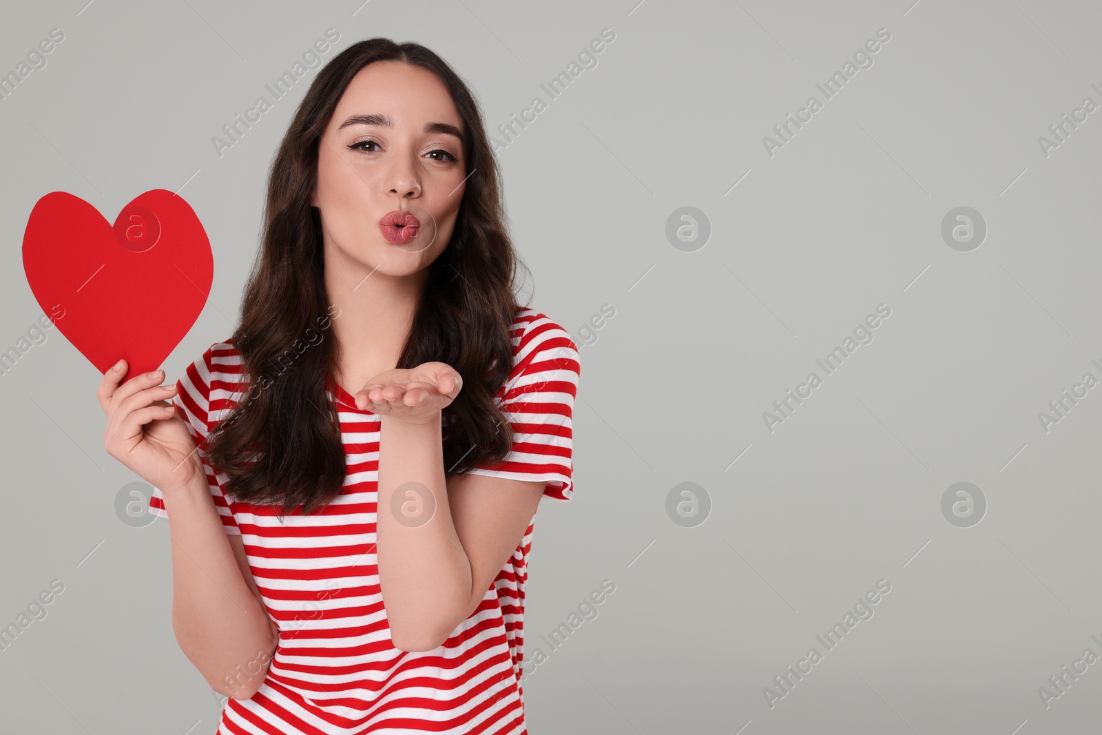 Photo of Beautiful young woman with paper heart blowing kiss on grey background, space for text