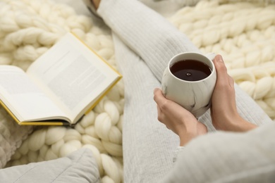 Woman with cup of tea and book sitting on soft plaid, closeup