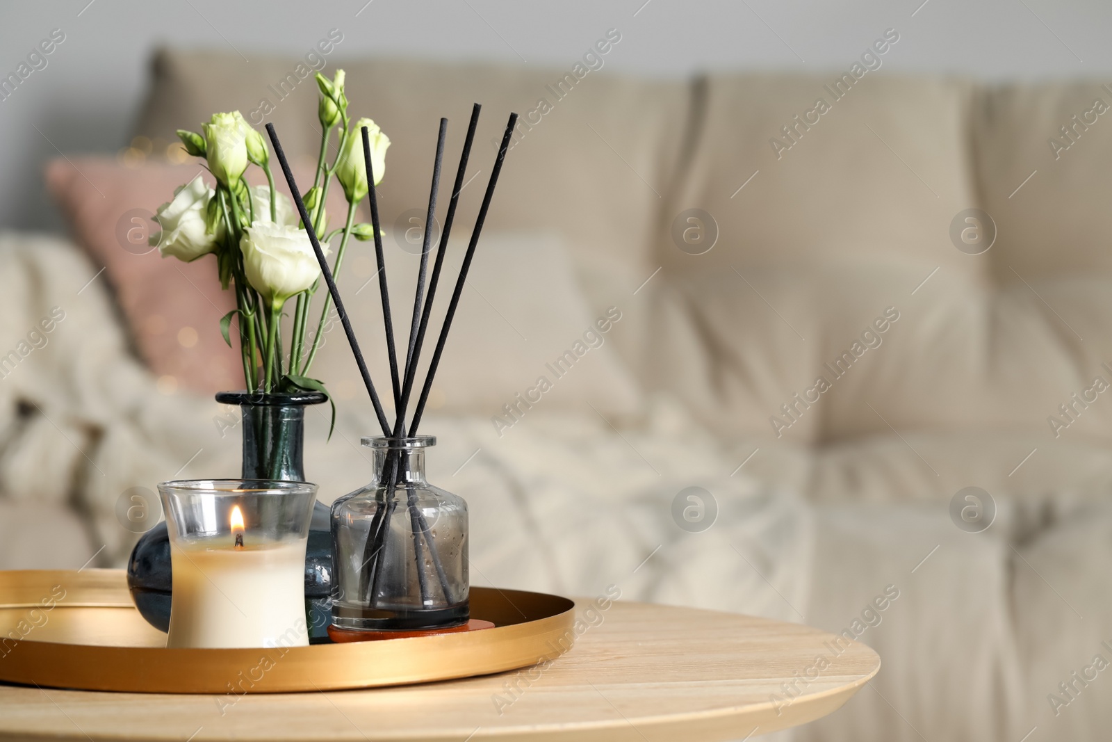 Photo of Burning candle with oil reed diffuser and bouquet on wooden table in living room