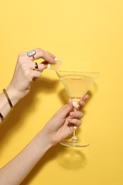 Photo of Woman adding lemon slice to martini glass of refreshing cocktail on yellow background, closeup