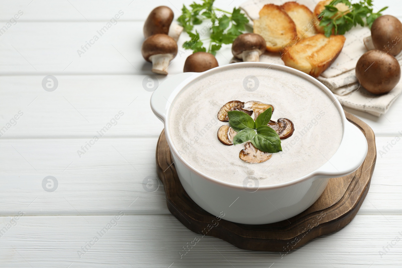 Photo of Delicious homemade mushroom soup in ceramic pot, croutons, fresh champignons and parsley on white wooden table. Space for text