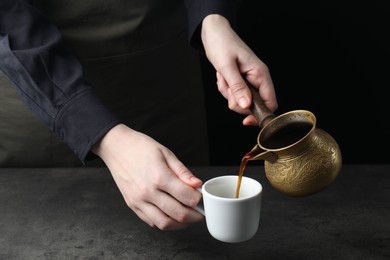 Photo of Turkish coffee. Woman pouring brewed beverage from cezve into cup at grey table, closeup