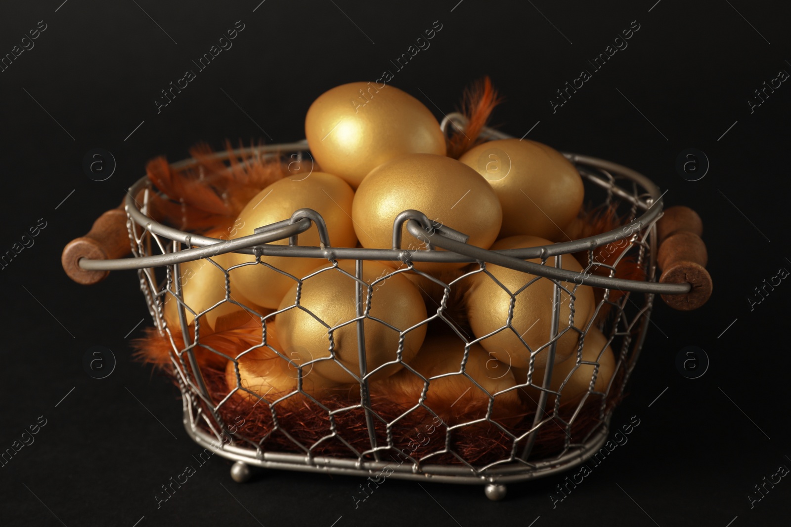 Photo of Shiny golden eggs with feathers in metal basket on black background