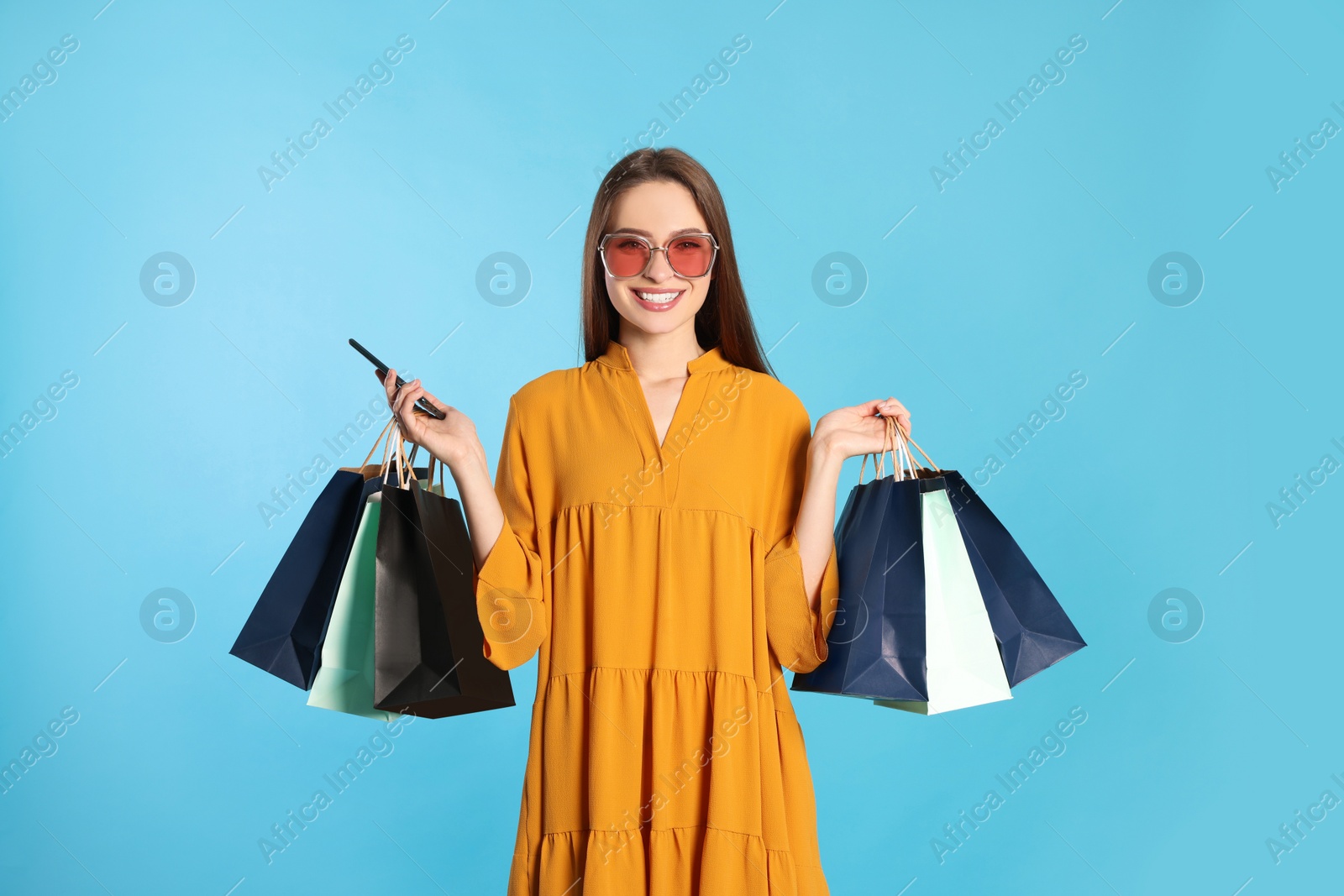 Photo of Happy young woman with shopping bags and smartphone on light blue background. Big sale