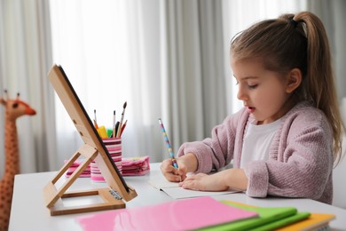 Adorable little girl doing homework with tablet at table indoors