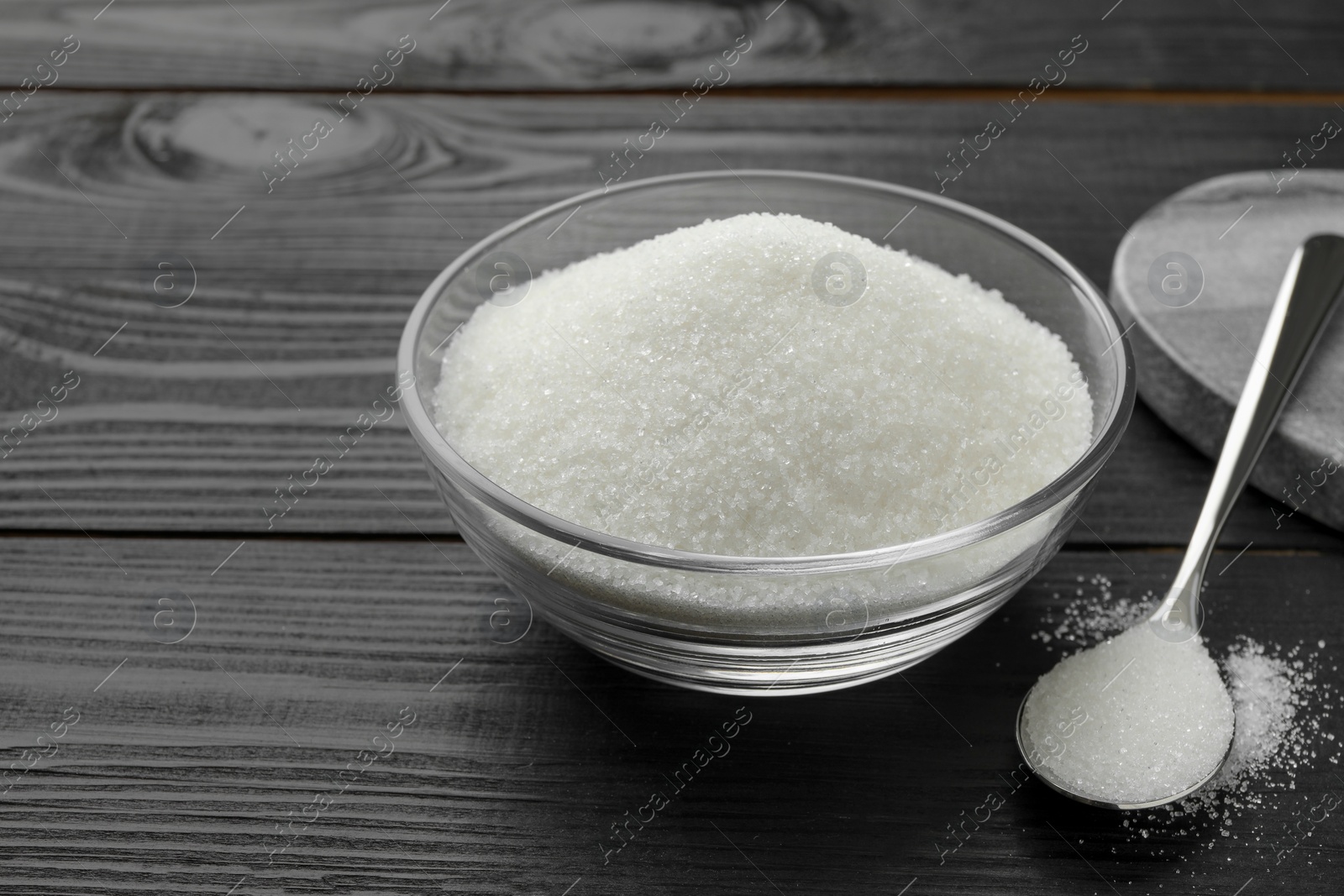 Photo of Granulated sugar in bowl and spoon on black wooden table, closeup. Space for text