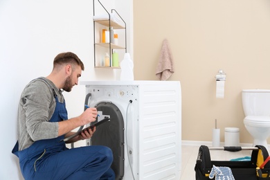 Photo of Young plumber with clipboard near washing machine in bathroom