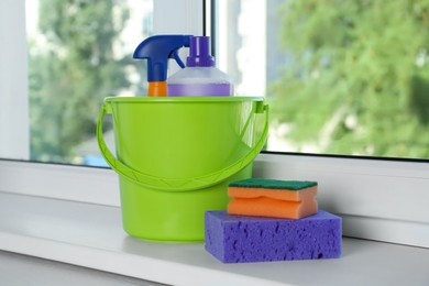 Photo of Green bucket with cleaning supplies and sponges on window sill indoors