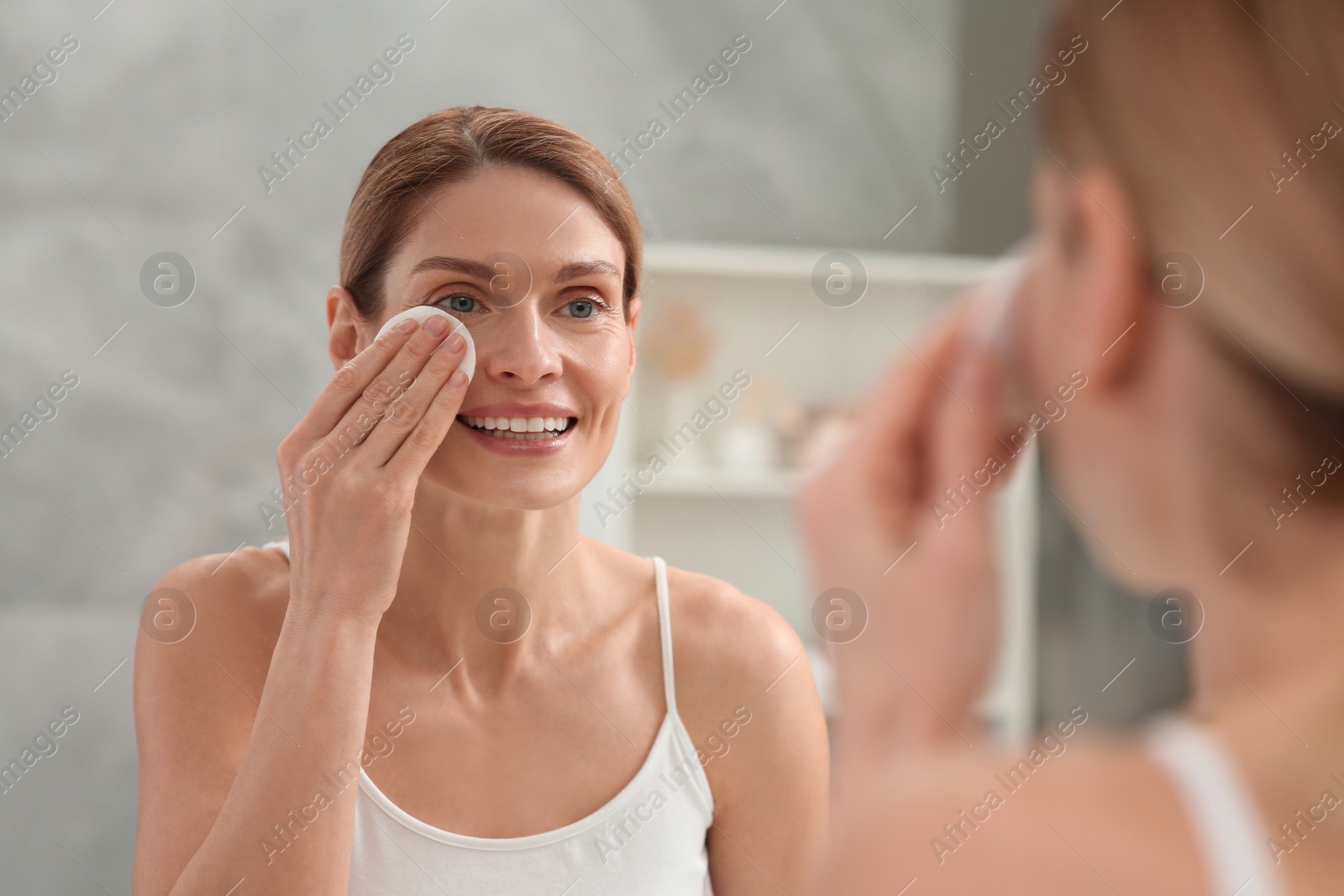 Photo of Beautiful woman removing makeup with cotton pad near mirror indoors