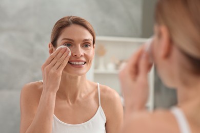 Beautiful woman removing makeup with cotton pad near mirror indoors