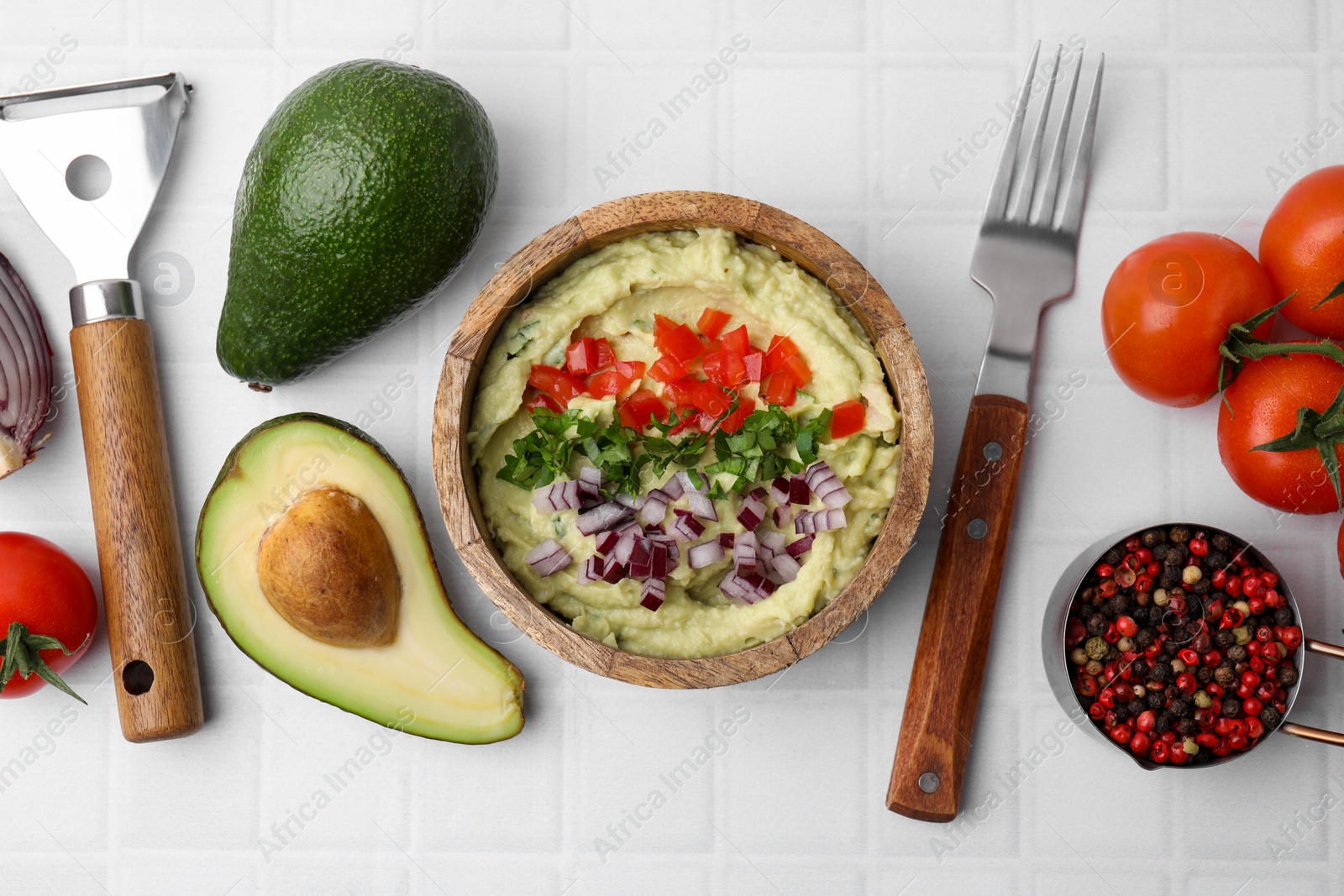 Photo of Bowl of delicious guacamole with onion, tomatoes and ingredients on white tiled table, flat lay