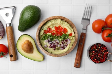 Bowl of delicious guacamole with onion, tomatoes and ingredients on white tiled table, flat lay