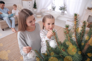 Photo of Mother with daughter decorating Christmas tree together while father and son sitting on sofa at home