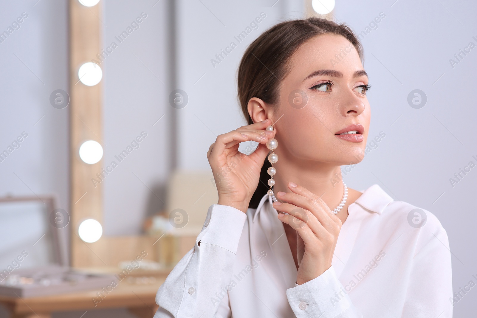 Photo of Young woman trying on elegant pearl earring indoors, space for text