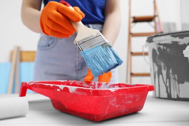 Photo of Woman taking light blue paint with brush from tray at white wooden table indoors, closeup