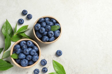 Photo of Tasty fresh blueberries with leaves in bowls on light grey table, flat lay. Space for text