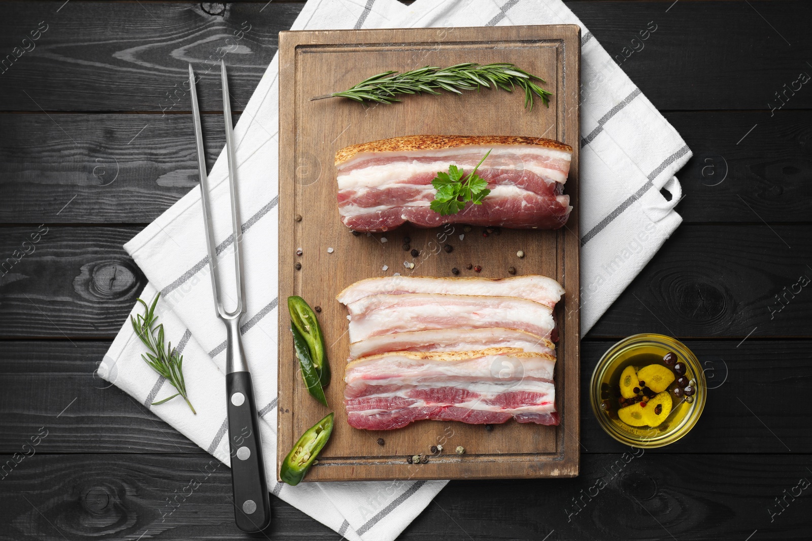 Photo of Pieces of raw pork belly, chili pepper, peppercorns, rosemary and parsley on black wooden table, flat lay