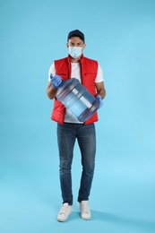Photo of Courier in face mask with bottle of cooler water on light blue background. Delivery during coronavirus quarantine