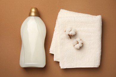 Photo of Bottle of fabric softener, terry towel and beautiful cotton flowers on brown background, flat lay