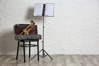 Photo of Trumpet, chair, case and note stand with music sheets near brick wall. Space for text