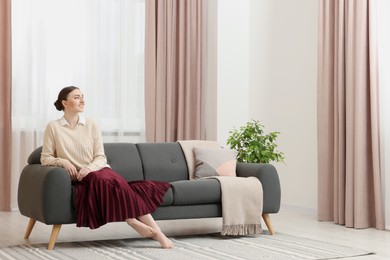 Woman sitting on sofa near window with stylish curtains at home. Space for text