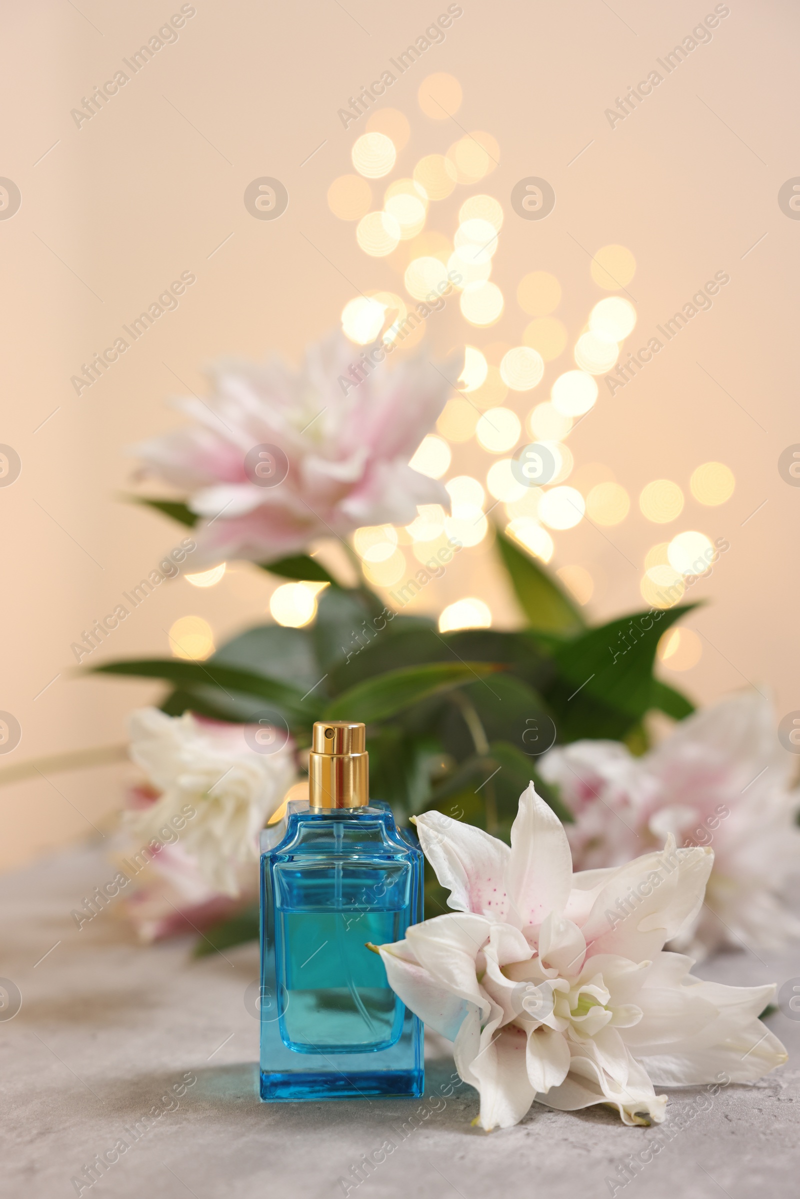 Photo of Bottle of perfume and beautiful lily flowers on table against beige background with blurred lights, closeup