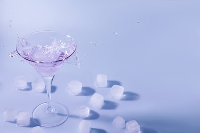 Photo of Cocktail splashing out of martini glass near ice cubes on light blue background, space for text