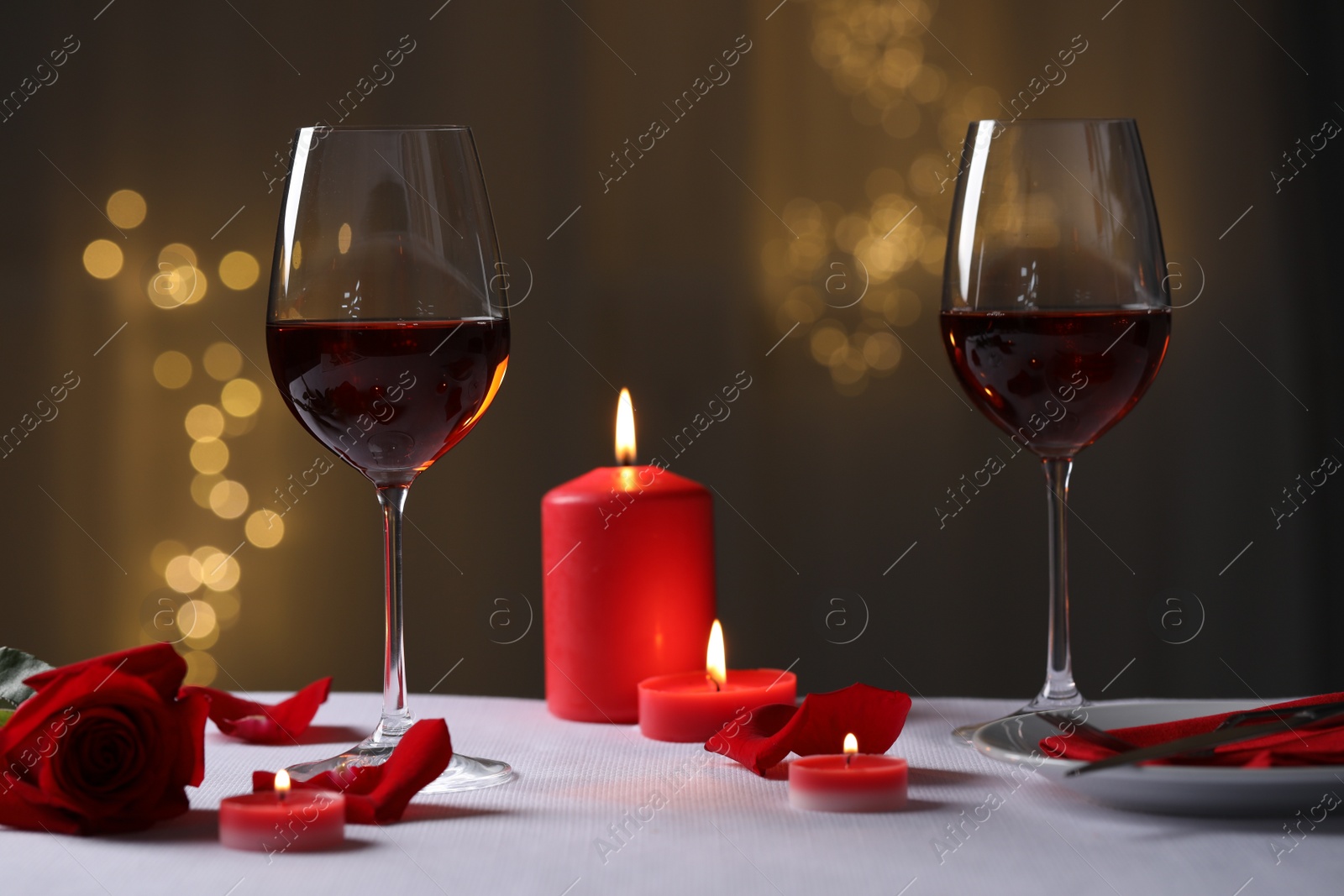 Photo of Glasses of red wine, burning candles and rose flower on grey table against blurred lights. Romantic atmosphere