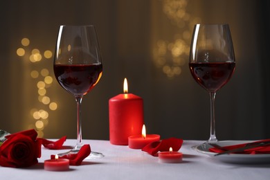 Glasses of red wine, burning candles and rose flower on grey table against blurred lights. Romantic atmosphere