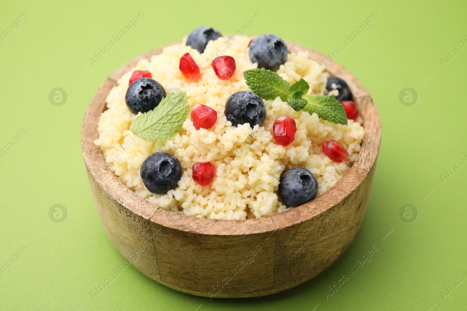 Photo of Bowl of tasty couscous with blueberries, pomegranate and mint on green background, closeup