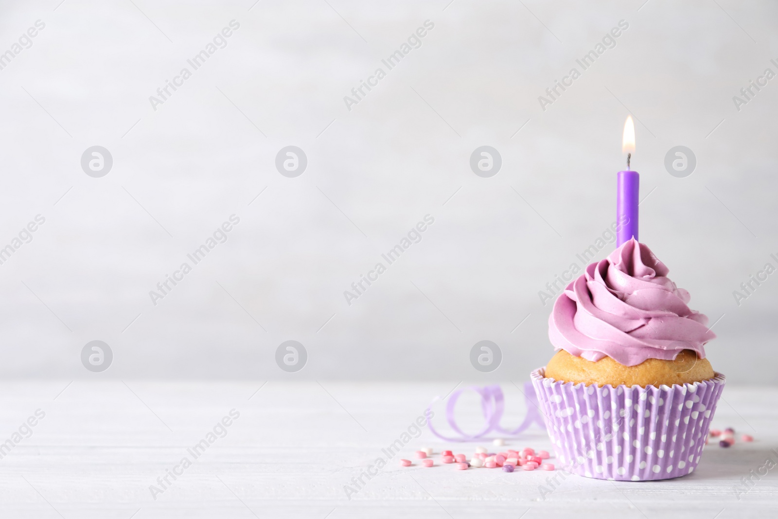 Photo of Delicious birthday cupcake with burning candle, sprinkles and streamer on white wooden table, space for text