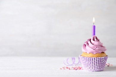 Delicious birthday cupcake with burning candle, sprinkles and streamer on white wooden table, space for text