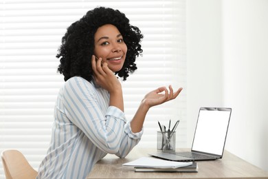 Happy young woman talking on phone while using laptop at wooden desk indoors