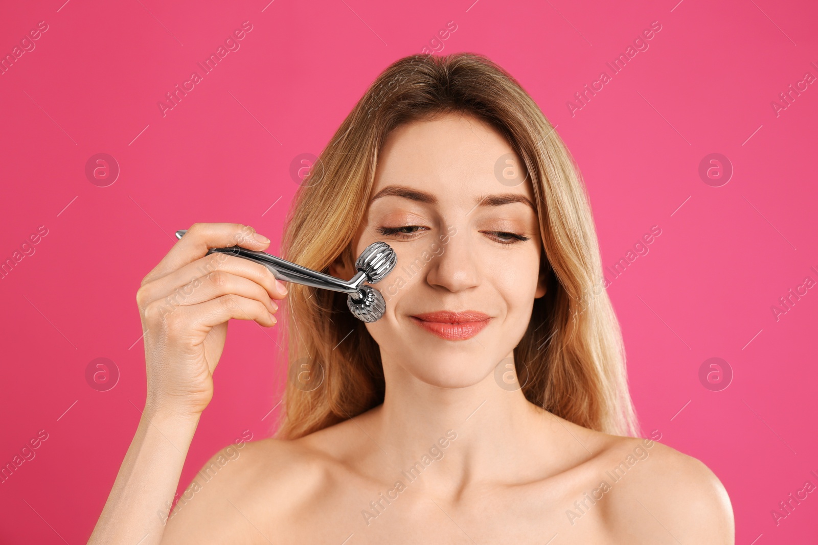 Photo of Young woman using metal face roller on pink background