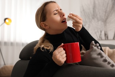 Sick woman with tissue and cup of drink sneezing on sofa at home. Cold symptoms