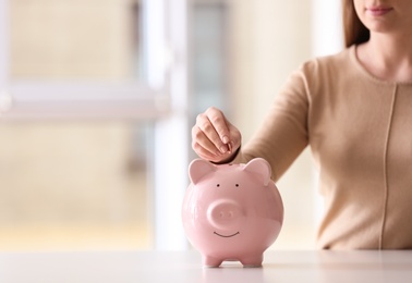 Photo of Woman putting coin into piggy bank at table indoors, closeup. Space for text