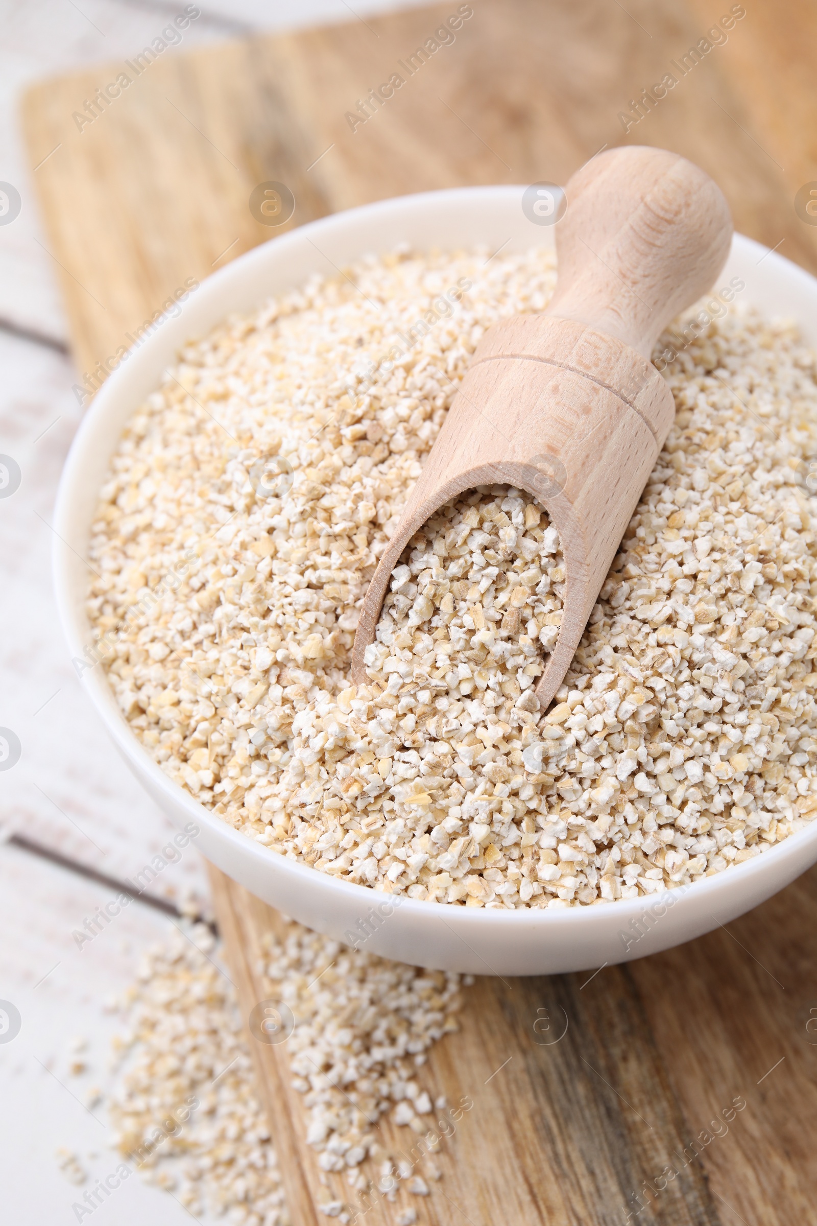 Photo of Raw barley groats and scoop in bowl on light wooden table, closeup