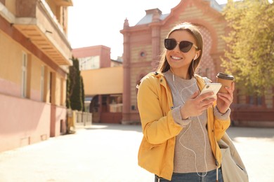 Happy young woman with coffee listening to music on city street in morning